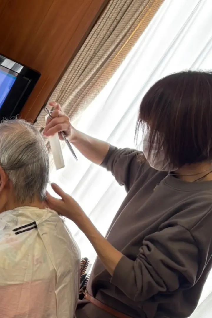 マスク姿の美容師が、椅子に座った高齢の方の髪を窓辺でカットしている様子